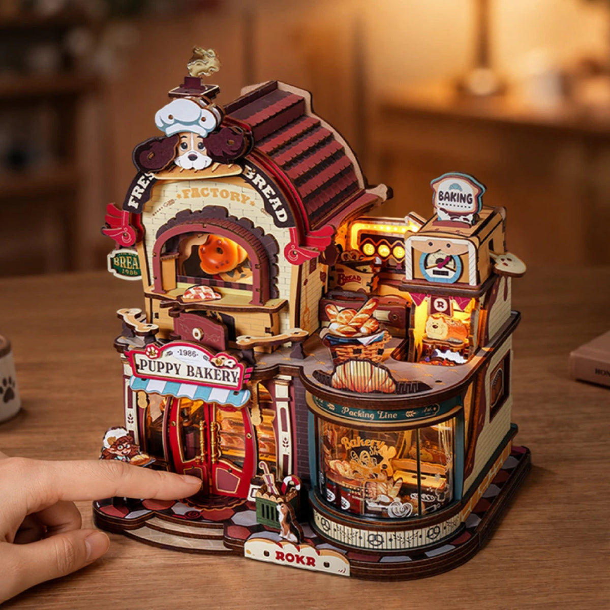 Dream Bread Factory 3D -Maquette en bois sur une table en bois, une main touche la boulangerie illuminée avec Puppy Bakery