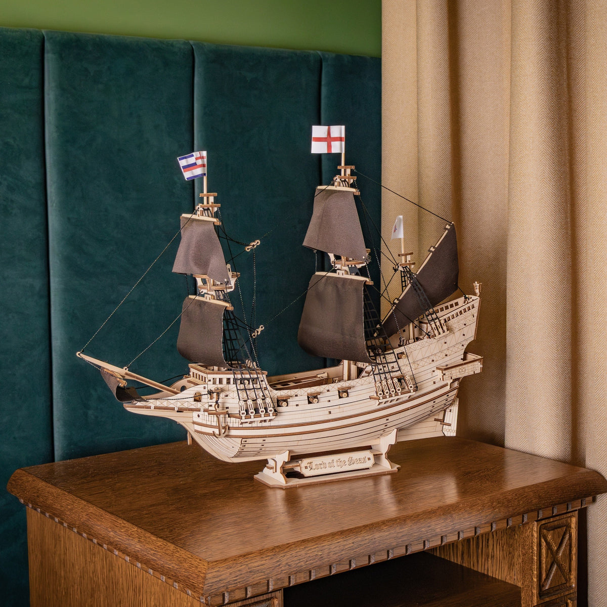 "Lord of the Seas" wooden model kit, a finished sailing ship on a wooden chest of drawers in front of a dark green velvet wall and curtain