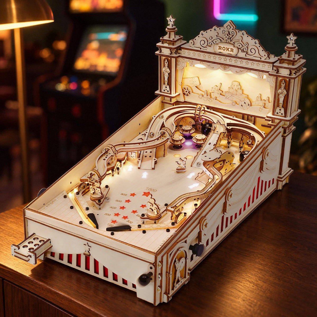 Une machine de flipper 3D en bois qui s'illumine le soir sur un buffet en noyer, avec derrière une borne d'arcade et une enseigne au néon