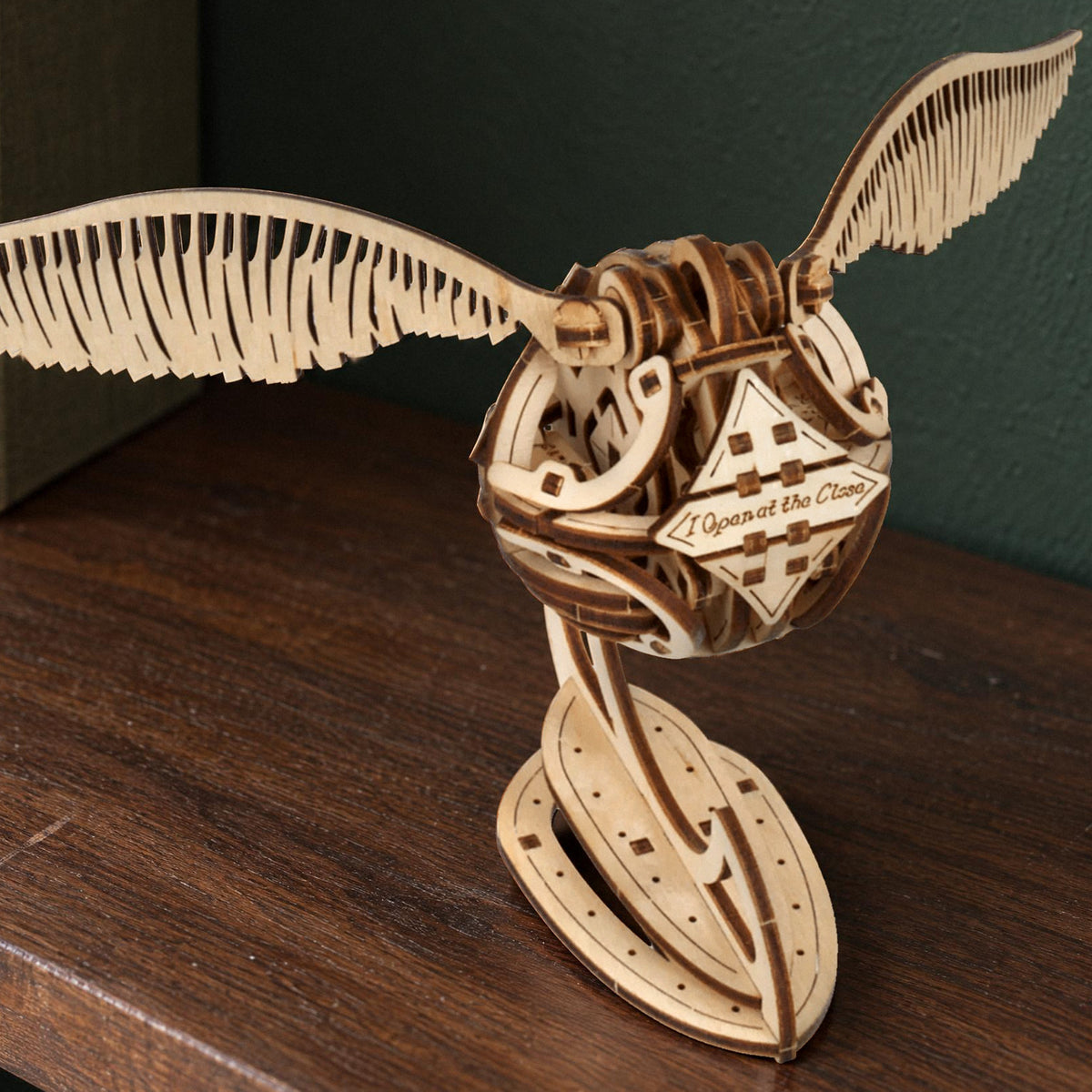 Golden Snitch – Harry Potter, Ugears wooden model kit on its stand on a shelf in front of a green wall in natural light