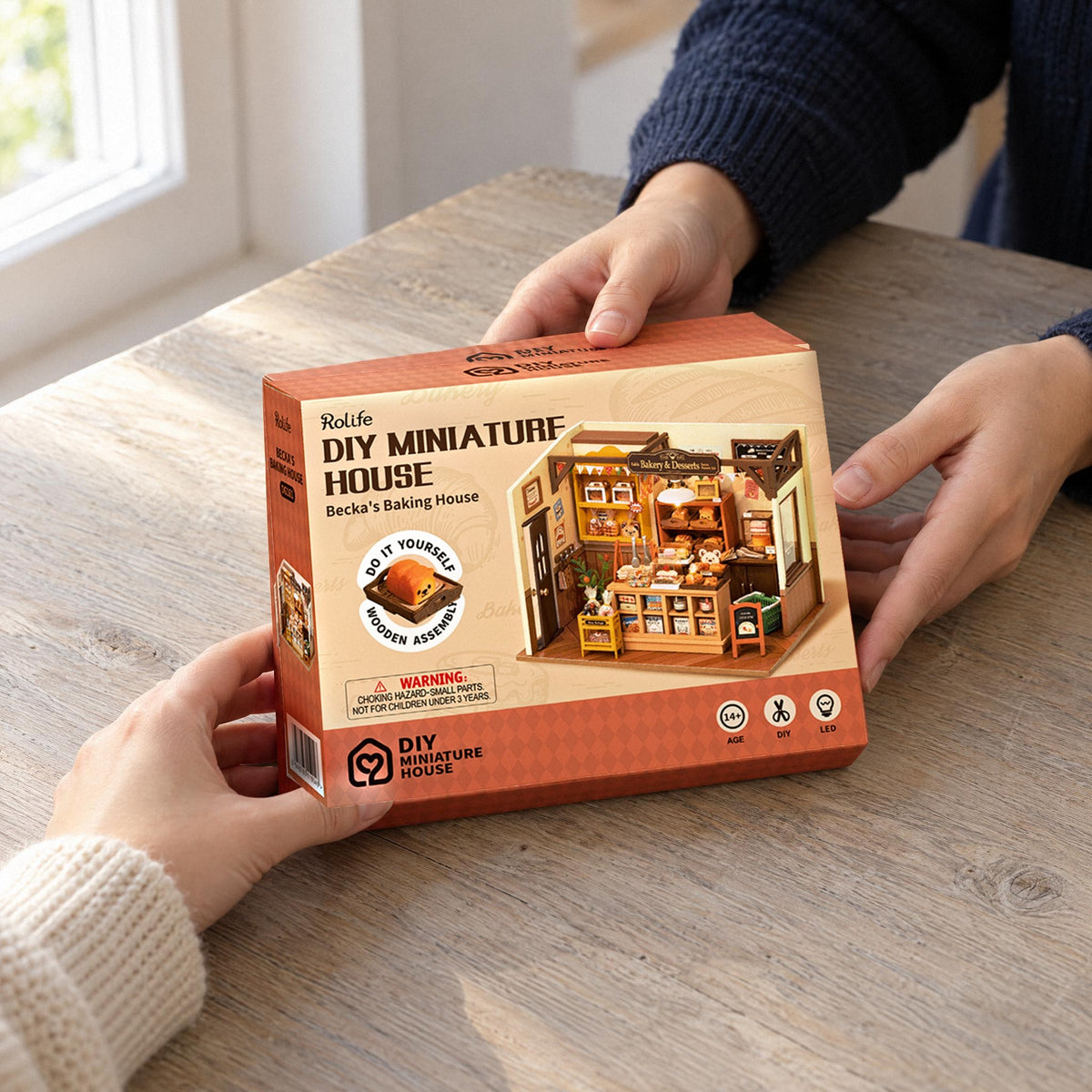 Becka’s Baking House (Bakery) Miniature House, Gift Box is handed to a second person at the dining table