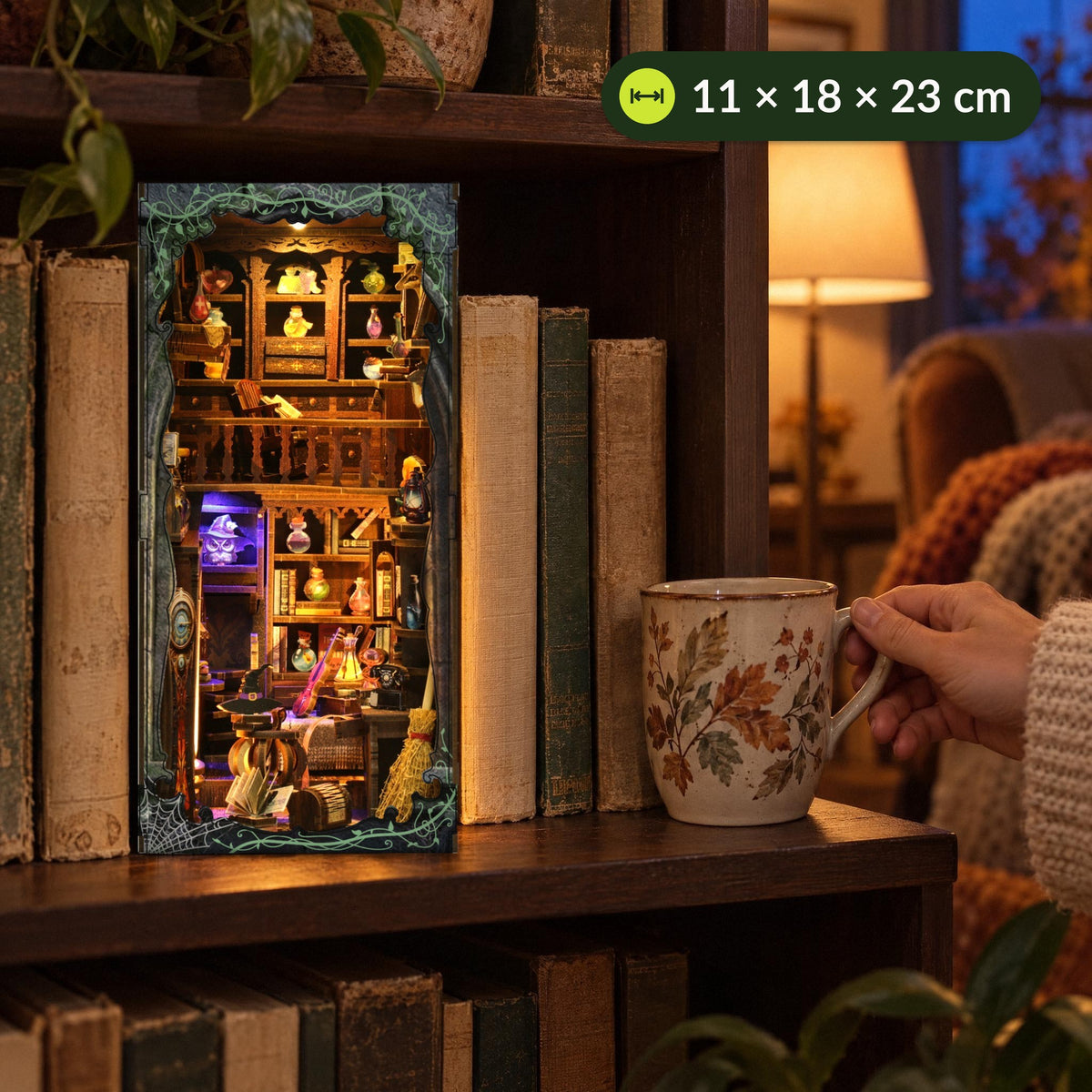 Magic Lab Book Nook, staat te schitteren op de plank in de woonkamer, met ernaast een hand met een theekopje, 11 × 18 × 23 cm
