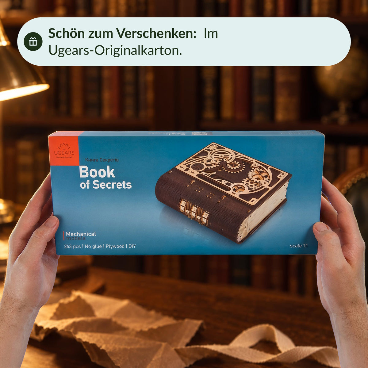 The original box for Ugears' "Book of Secrets" is presented with both hands, with wrapping paper and a ribbon behind it