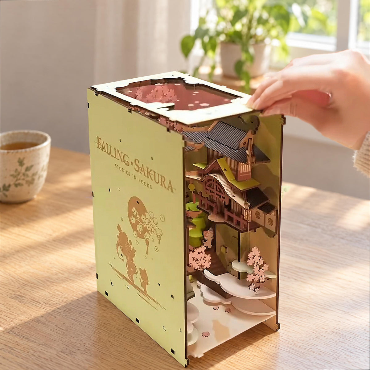 Falling Sakura Book Nook, linke Seitenwand wie ein Buchdeckel graviert, eine Hand setzt das Dach auf