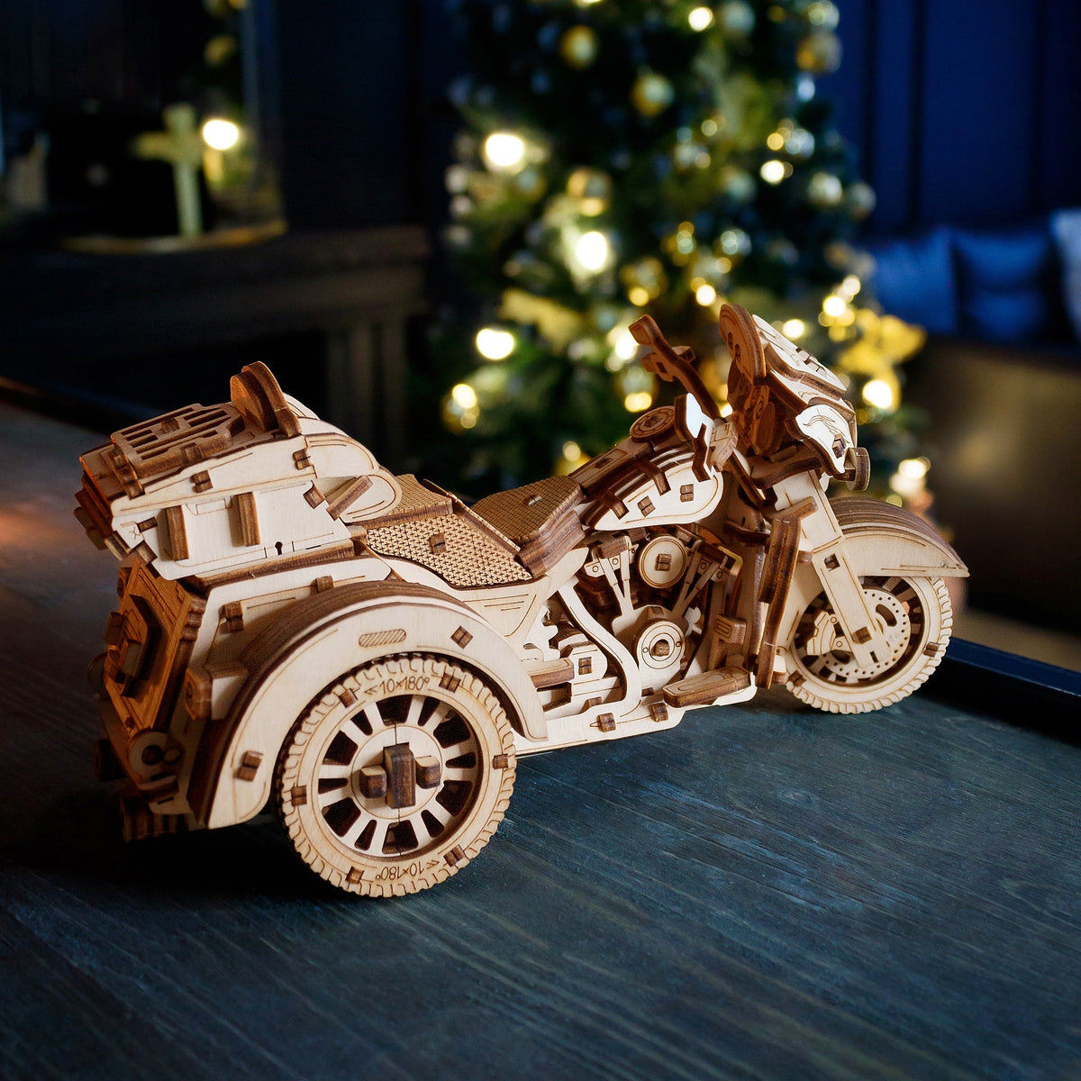 Un trike en bois, sous forme de kit mécanique, posé sur une table en bois devant le sapin de Noël illuminé, le soir