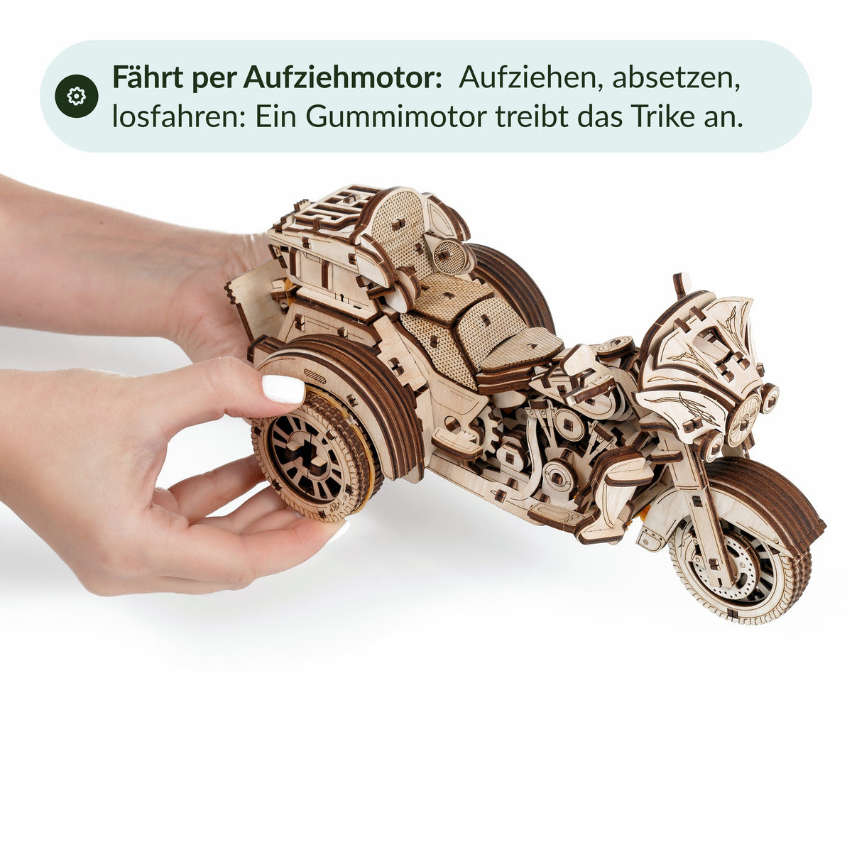 Hände ziehen das Holz-Trike am Hinterrad auf, ein Gummimotor treibt das Modell an